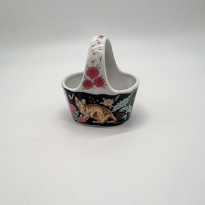 May include: A small ceramic basket with a high arched handle, featuring a black exterior decorated with a brown tabby cat, pink flowers, and green foliage. The handle is white with a delicate floral pattern of pink roses and small white blossoms.