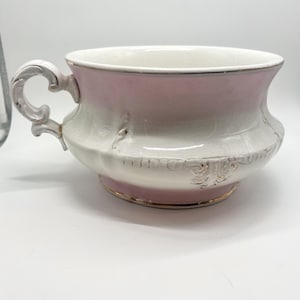 May include: A decorative ceramic bowl with a handle, featuring a pink and white color scheme. The bowl has a gold trim and ornate detailing around the base and handle. The handle is curved and detailed.