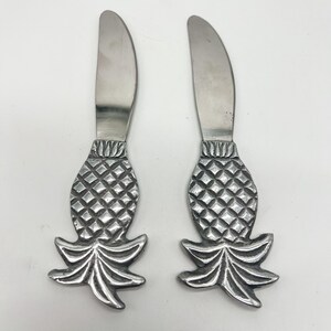 May include: Two silver-toned metal knives featuring handles shaped like pineapples. Each knife has a smooth, curved blade and a textured, diamond-patterned pineapple body with a flared, leafy base at the bottom of the handle.