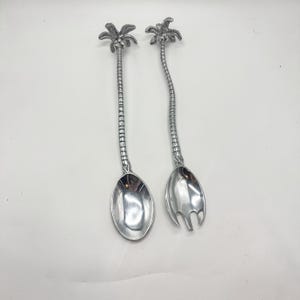 May include: Two silver-colored serving utensils. One is a spoon, and the other is a fork. Both have long, textured handles and palm tree designs at the top. The spoon and fork are designed for serving food.