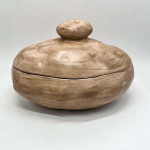 May include: A beige ceramic lidded container with a rounded, organic shape. The lid has a small, knob-like handle. The surface has a textured, wood-like appearance, with subtle variations in color. The container is likely for storage or decorative use.