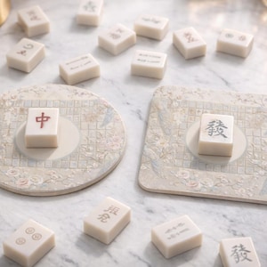 Könnte beinhalten: Eine Sammlung elfenbeinfarbener Mahjong-Steine, die um zwei dekorative Untersetzer herum verstreut sind. Die Untersetzer sind beige mit Blumen- und Vogelmustern. Jeder Stein hat ein einzigartiges Symbol oder Zeichen in Schwarz oder Rot.
