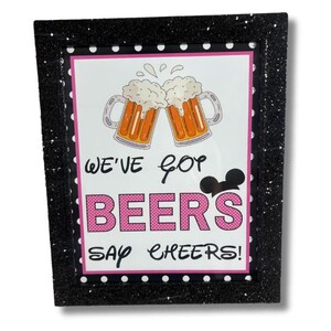Könnte beinhalten: Ein schwarzer gerahmtes Bild mit weißem Hintergrund und rosa Punkten. Das Bild zeigt zwei Cartoon-Bierkrüge, die anstoßen, mit dem Text "We've got beers say cheers!" und einem Mickey-Maus-Kopf.