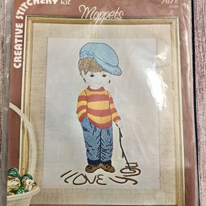 May include: A needlepoint kit featuring a boy with a blue hat and red and yellow striped shirt holding a stick. The boy is standing in front of a white background with the words "I LOVE YOU" written in black. The kit includes stamped fabric, yarn, and instructions.