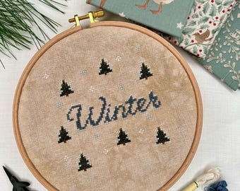 Può includere: Un telaio da ricamo con la parola "Winter" in filo blu, circondata da piccoli disegni di alberi verdi. Il telaio è posto su un tessuto beige, con un paio di forbici nere e rocchetti di filo nelle vicinanze. Tessuto decorativo con motivi invernali sullo sfondo.