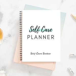 May include: A white spiral bound notebook with the text "Self Care PLANNER" and "Self Care Seeker" on the cover. The notebook is on a pink and blue background.