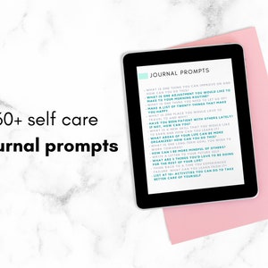 May include: A tablet displaying a list of 60+ self-care journal prompts on a pink surface. The text on the tablet is in teal and white. The tablet is next to a black pen.