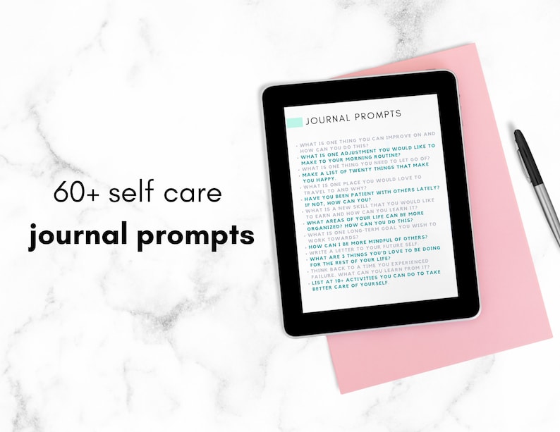 May include: A tablet displaying a list of 60+ self-care journal prompts on a pink surface. The text on the tablet is in teal and white. The tablet is next to a black pen.