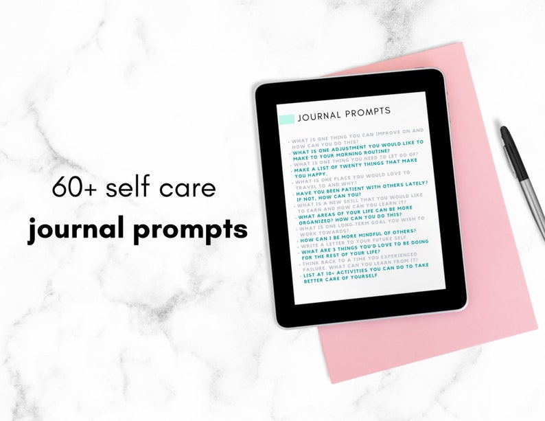 May include: A tablet displaying a list of 60+ self-care journal prompts on a pink surface. The text on the tablet is in teal and white. The tablet is next to a black pen.