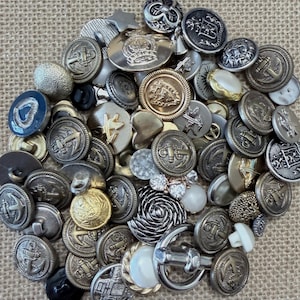 May include: A selection of vintage buttons in diverse shapes and metallic finishes. The buttons feature anchor designs, crown emblems, and decorative elements. Colours include silver, gold, and pearl. Suitable for sewing, crafting, or collecting.