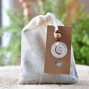 May include: A small off-white drawstring fabric pouch with a brown rectangular cardboard tag attached by a white string. The tag features a circular logo with a crescent moon and floral illustration, the text Yarrow Moon, and a heart-shaped cutout.