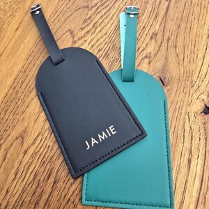 May include: Two luggage tags, one black and one teal, rest on a wooden surface. The black tag has the name "JAMIE" printed in white. Both tags have a matching black strap with a silver buckle.