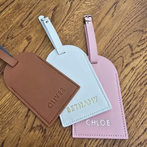May include: Three personalized luggage tags in brown, white, and pink. Each tag has a different name embossed in gold or silver lettering: Oliver, Bethany, and Chloe. The tags have matching straps and silver or gold buckles.