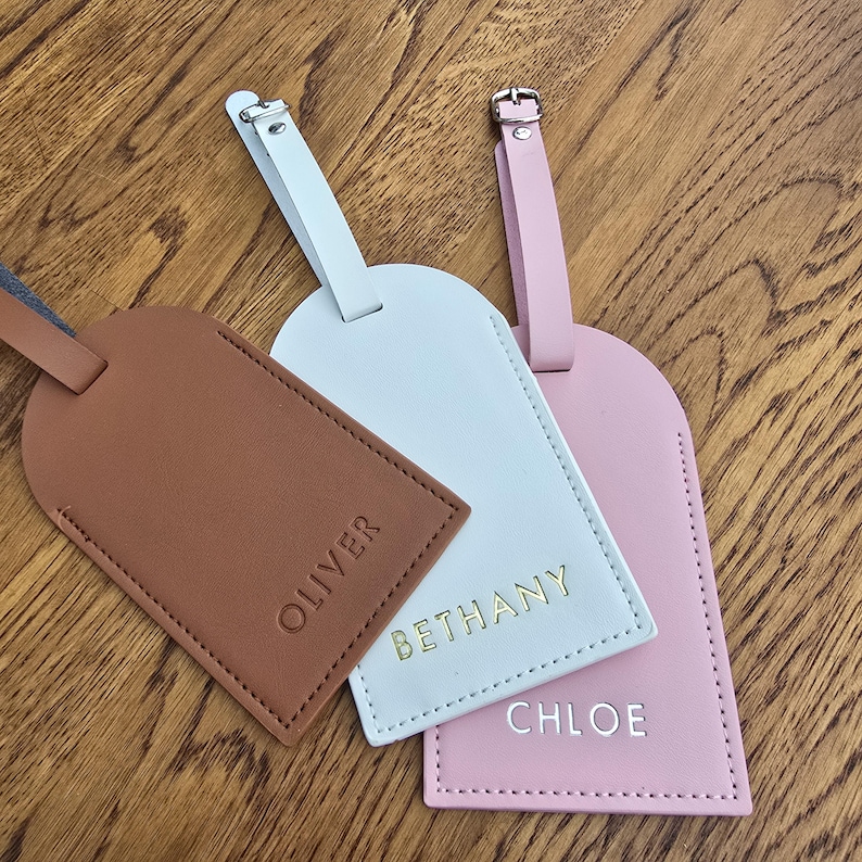 May include: Three personalized luggage tags in brown, white, and pink. Each tag has a different name embossed in gold or silver lettering: Oliver, Bethany, and Chloe. The tags have matching straps and silver or gold buckles.