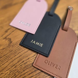 May include: Three personalized luggage tags in pink, black, and brown leather. Each tag has a name embossed in a contrasting color: "Chloe", "Jamie", and "Oliver". The tags are rectangular with a curved top and a strap.