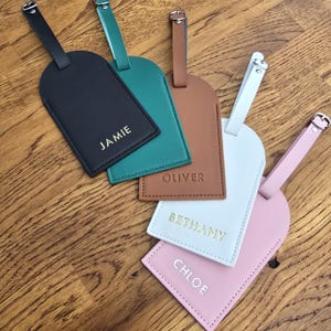 May include: A collection of personalized luggage tags in various colors, including black, teal, brown, white, and pink. Each tag features a name in gold lettering. The tags have a buckle closure and a strap.