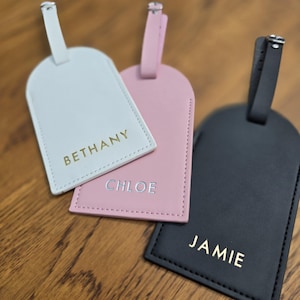 May include: Three personalized luggage tags in white, pink, and black. Each tag has a silver metal loop and a name printed in a contrasting color: "Bethany" in gold, "Chloe" in silver, and "Jamie" in gold. The tags are made of a smooth material.