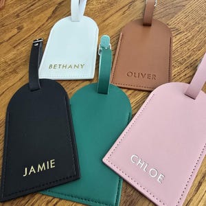 May include: Five personalized luggage tags in various colors: white, brown, black, green, and pink. Each tag features a name in a contrasting color: Bethany, Oliver, Jamie, and Chloe. The tags have a stitched border and a strap with a buckle.