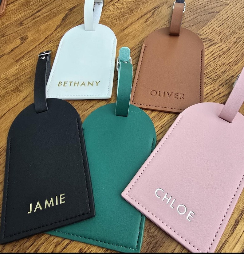 May include: Five personalized luggage tags in various colors: white, brown, black, green, and pink. Each tag features a name in a contrasting color: Bethany, Oliver, Jamie, and Chloe. The tags have a stitched border and a strap with a buckle.