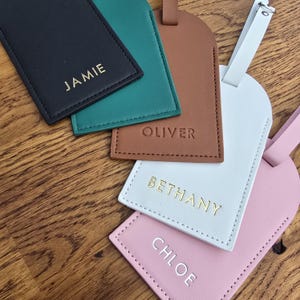 May include: A collection of personalized luggage tags in various colors, including black, teal, brown, white, and pink. Each tag features a name in gold lettering: Jamie, Oliver, Bethany, and Chloe. The tags are made of a leather-like material.