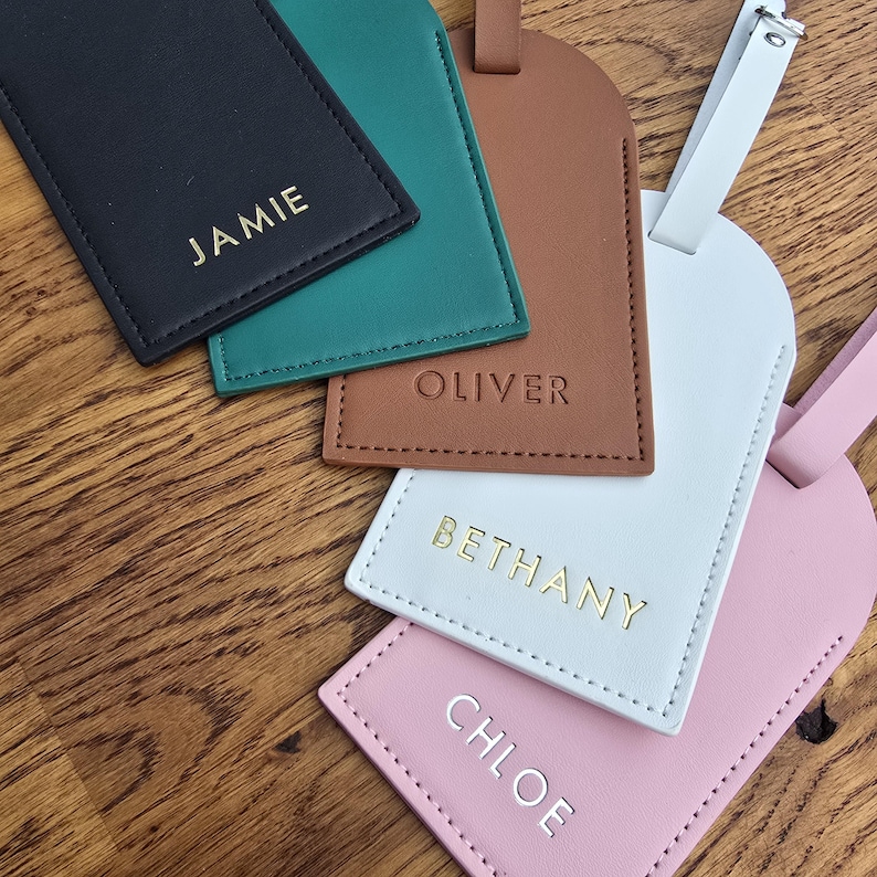 May include: A collection of personalized luggage tags in various colors, including black, teal, brown, white, and pink. Each tag features a name in gold lettering: Jamie, Oliver, Bethany, and Chloe. The tags are made of a leather-like material.