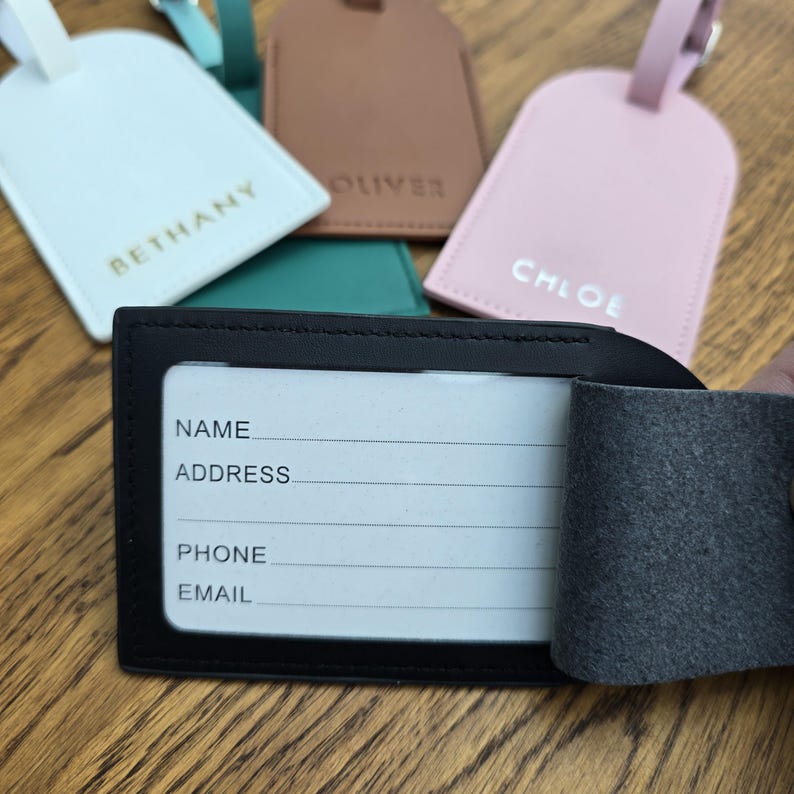 May include: A collection of personalized luggage tags in various colors, including white, brown, and pink. Each tag features a name in a contrasting color. A black luggage tag with a clear window for contact information is in the foreground.