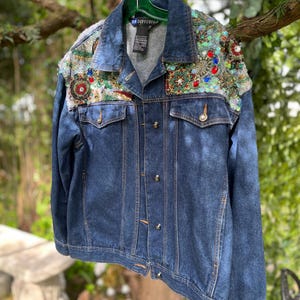 May include: A dark blue denim jacket with decorative embellishments on the shoulders. The jacket has a button-up front, two chest pockets, and a collar. The embellishments include colorful sequins and beads. The jacket is hanging from a green hanger.