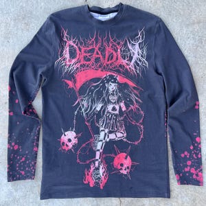 May include: Dark gray long-sleeve shirt with a graphic of a gothic anime girl holding a scythe, surrounded by skulls and flames. The word "DEADLY" is printed across the top in pink and white. The sleeves have pink splatter details.
