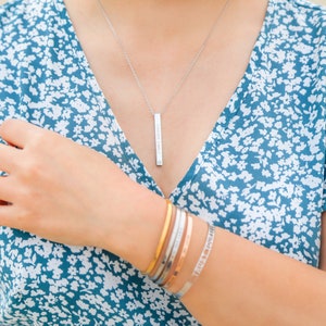 Op de afbeelding: Een zilveren hangerketting met de woorden "Forever Family" gegraveerd. De ketting wordt gecombineerd met verschillende armbanden in goud, zilver en roségoud. De armbanden hebben gegraveerde tekst. De persoon draagt een blauwe bloemenjurk.