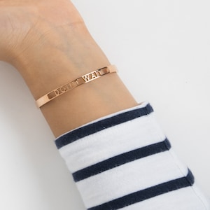 May include: Rose gold cuff bracelet with the engraved words "DON'T WAIT". The bracelet is a simple, elegant design. The person is wearing a striped shirt with navy blue and white stripes.