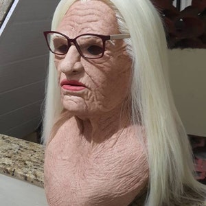 Puede incluir: Una m&aacute;scara de l&aacute;tex de aspecto realista de una persona mayor con cabello blanco y arrugas. La m&aacute;scara lleva gafas con montura roja.