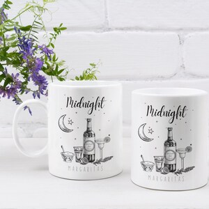 May include: Two white ceramic mugs with black text and illustrations. The mugs feature a crescent moon, a star, a bottle of tequila, a lime wedge, a salt shaker, a shot glass, and a margarita glass. The text on the mugs reads "Midnight Margaritas".