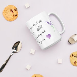 May include: White ceramic mug with a black and purple design. The mug has the text "Toss your left shoulder a sprinkle of lavender for luck whenever you can." with a small drawing of a salt shaker, lavender flowers, and a heart.