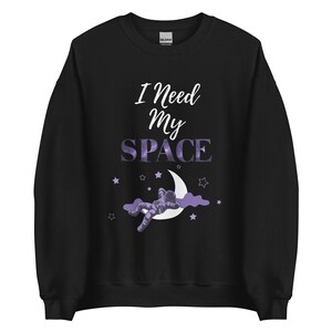 Könnte beinhalten: Schwarzer Pullover mit einer lila und weißen Grafik eines Astronauten, der auf einer Mondsichel mit Sternen schläft. Der Text "I Need My Space" ist in Weiß gedruckt.