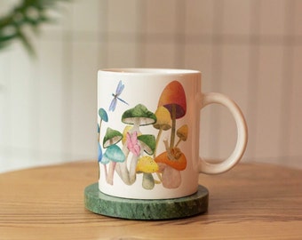 May include: White ceramic mug featuring a colorful watercolor illustration of various mushrooms and a dragonfly. The mug is resting on a green marble coaster. The mug has a curved handle and is suitable for hot or cold beverages.