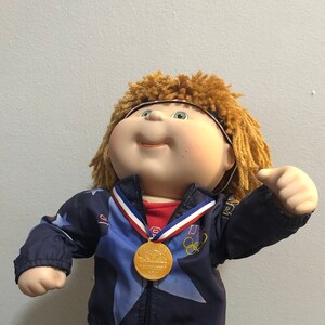 May include: A Cabbage Patch Kid doll wearing a blue and red tracksuit with stars and the words "Olympic Games" and "USA". The doll is holding a gold medal and has a red, white, and blue ribbon around its neck.