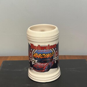 May include: Cream-colored ceramic beer stein featuring the Budweiser Racing logo with a race car graphic. The stein has a cylindrical shape with a wide opening and a decorative design around the top and bottom.