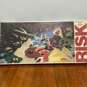 May include: Vintage Risk board game box featuring a world map with territories and game pieces. The box has the title "Risk" in large red letters and the words "Parker Brothers World Conquest Game". The game board shows a cannon.