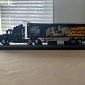 May include: A black toy semi-truck with a white trailer. The trailer has a yellow and red design with the words "MAC TOOLS Racing" and "Havoline". The truck is a model of a real-life semi-truck.
