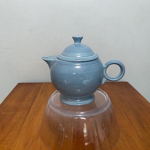 May include: A blue ceramic teapot with a lid, spout, and handle. The teapot is round and sits on a clear glass bowl. The teapot is a light blue color and appears to be made of ceramic.