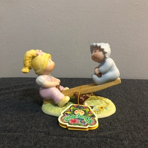 May include: A porcelain figurine of two Cabbage Patch Kids on a seesaw. The girl is wearing a pink dress and the boy is wearing a blue outfit. The figurine is on a green base with flowers and the Cabbage Patch Kids logo.