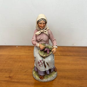 May include: A ceramic figurine of a seated elderly person wearing a headscarf and a pink blouse. The figure holds a basket of purple grapes. The skirt is white with brown patches. The figurine is on a green base.