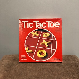 May include: A red box with the words "Tic Tac Toe" in white letters. The box contains a game board with yellow X's and O's. The box also includes the words "Pressman" in white letters on the bottom right corner.