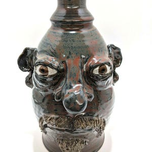 May include: A ceramic vase with a face design. The vase is primarily brown with blue and red accents. It features a detailed face with eyes, a nose, a mustache, and ears. The vase has a narrow neck and a wide base.