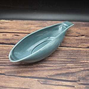 May include: A boat-shaped ceramic dish with a smooth, glossy finish in a muted blue-green color. The vessel features an elongated, tapered design with curved sides and a slightly raised, pointed end, creating a deep, open basin for serving.