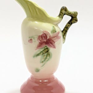 May include: A vintage ceramic pitcher with a floral design. The pitcher features a light green spout and handle, a pink base, and a cream-colored body adorned with a red flower and green leaves. The handle has a textured, branch-like appearance.