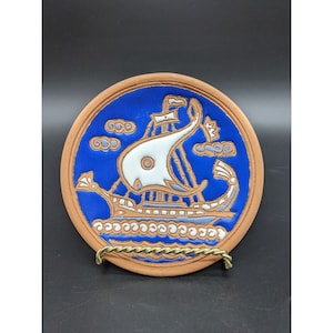 May include: A round terracotta plate with a blue and white painted design of a ship sailing on the sea. The ship has a white sail and a blue hull. The sea is depicted with white waves.