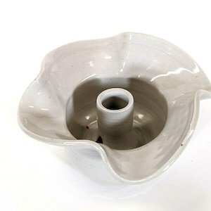 May include: A light gray ceramic candle holder with a unique, wavy rim design. The center features a cylindrical holder for a single candle. The overall shape is reminiscent of a flower.