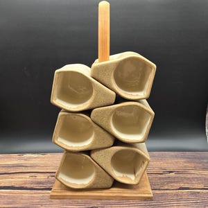 May include: A set of six beige ceramic cups with a speckled texture and smooth interior, stacked in two columns on a wooden base. A vertical wooden handle extends from the center of the base, providing a central support for the stacked ceramic cups.