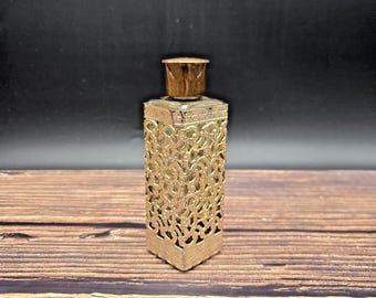 May include: A gold-colored perfume bottle with an ornate, openwork design. The bottle has a rectangular shape with a matching cap. The intricate pattern covers the sides of the bottle, creating a vintage or antique aesthetic. The bottle is displayed on a wooden surface.
